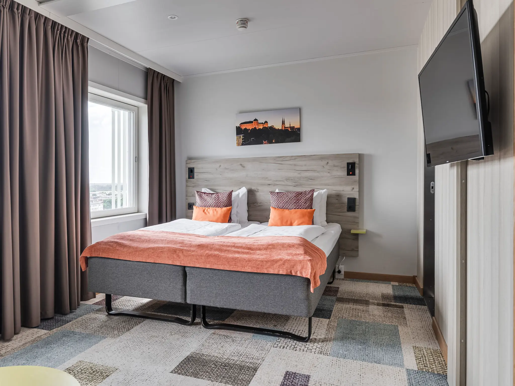 Modernt hotellrum med dubbelsäng, grå sänggavel och ljusorange accenter. Stort fönster med mörka gardiner och platt-TV.