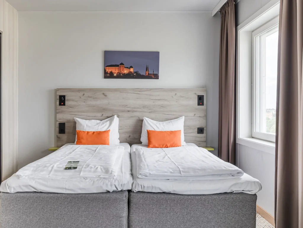 Cozy hotel room with a double bed, white linens and pillows in bright orange. A photo of Uppsala hangs above the bed and next to it is a large window with brown curtains.