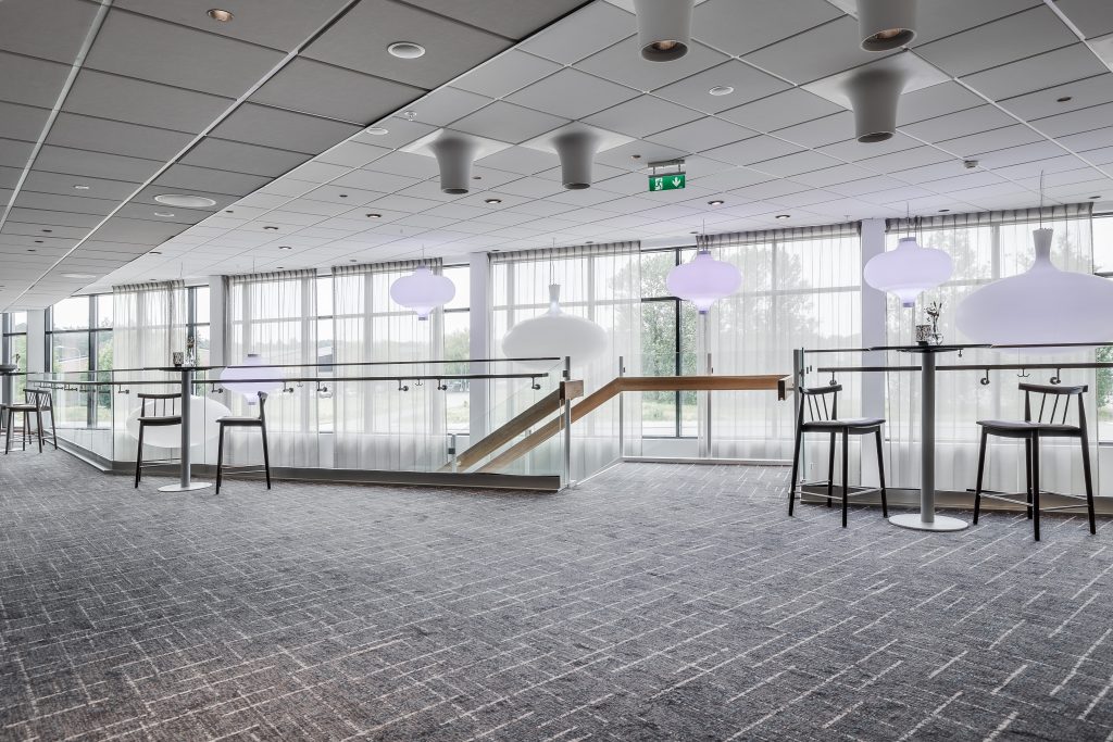 Modern loungedel med grå heltäckningsmatta, höga bord och svarta pallar. Stora fönster med skira gardiner och stora taklampor