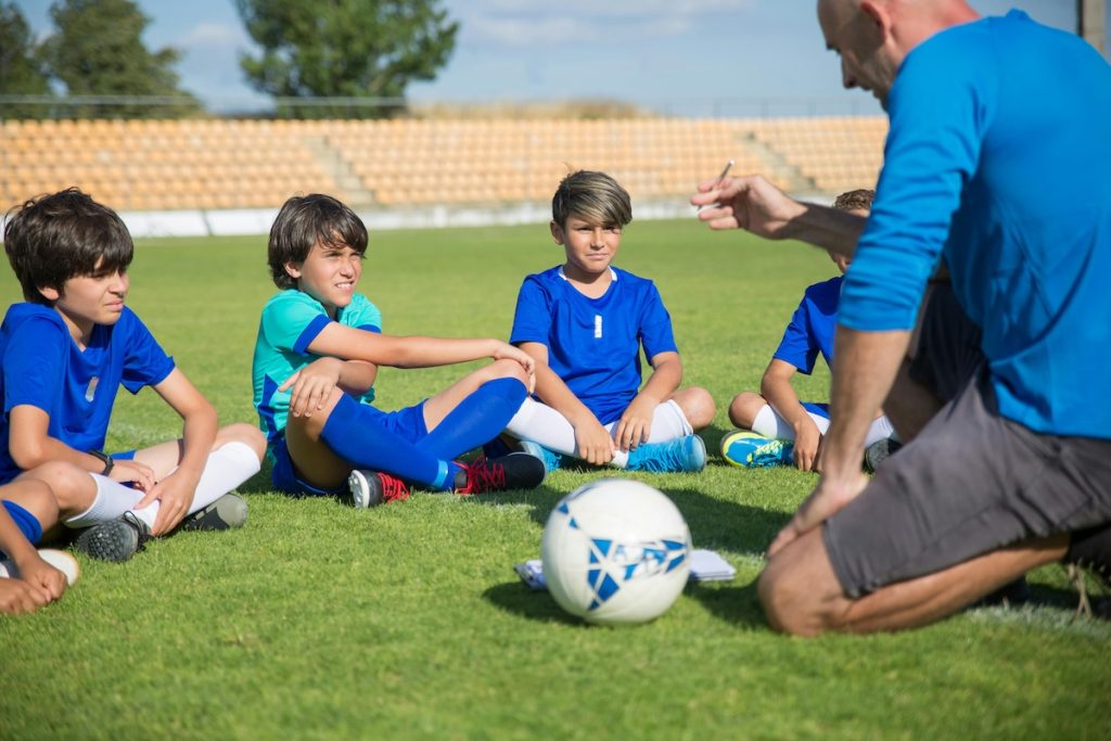 En fotbollstränare knäböjer på gräset och instruerar en grupp unga pojkar i blå uniformer som sitter i en cirkel och förmedlar lagarbete och fokus.